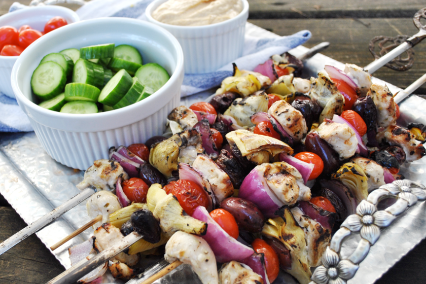 Grilled Greek chicken kabobs with cherry tomatoes, red onions, artichoke hearts, and olives on metal skewers. Served with sides of sliced cucumbers, cherry tomatoes, and a bowl of hummus.