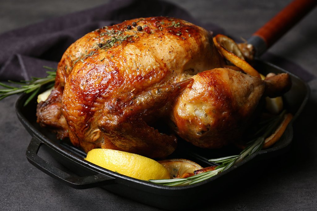 Perfectly roasted whole chicken seasoned with rosemary and lemon, presented in a cast iron skillet. The golden-brown skin is crispy and flavorful, accompanied by lemon wedges and fresh rosemary sprigs.