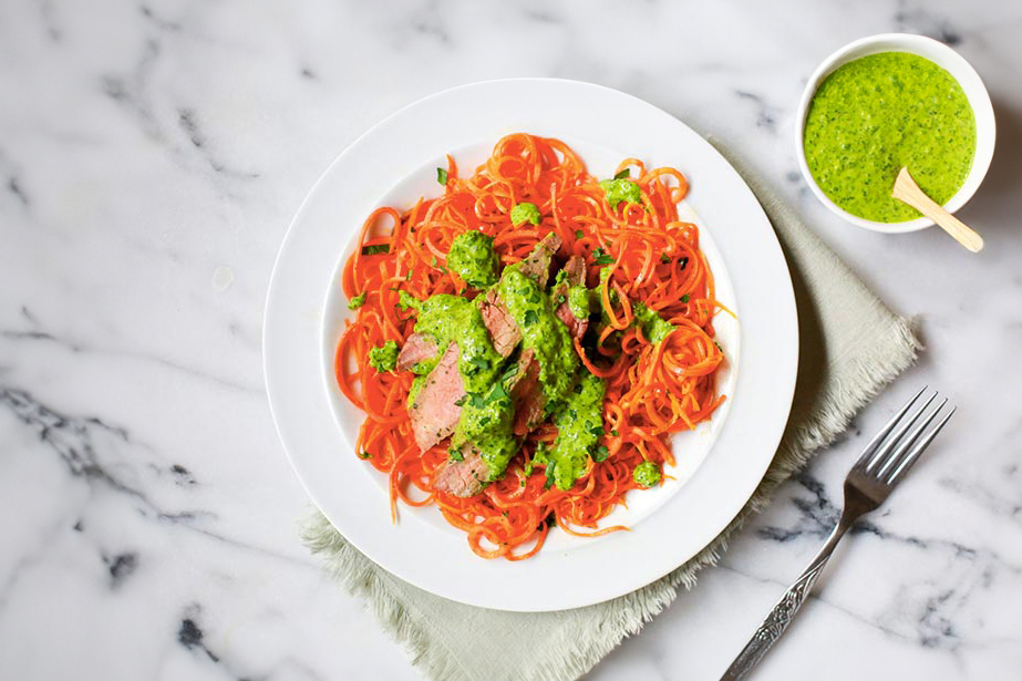 A plate of spiralized carrot noodles topped with sliced flank steak and drizzled with a vibrant green chimichurri sauce. The dish is accompanied by a small bowl of extra chimichurri sauce, set on a marble surface with a fork nearby.