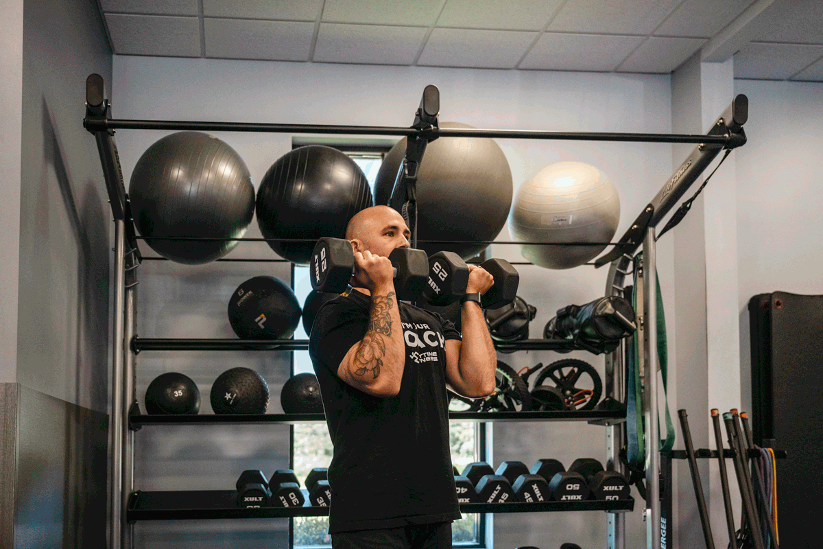 Anytime Fitness coach performing Arnold press exercise with dumbbells in a gym, demonstrating proper shoulder workout technique.