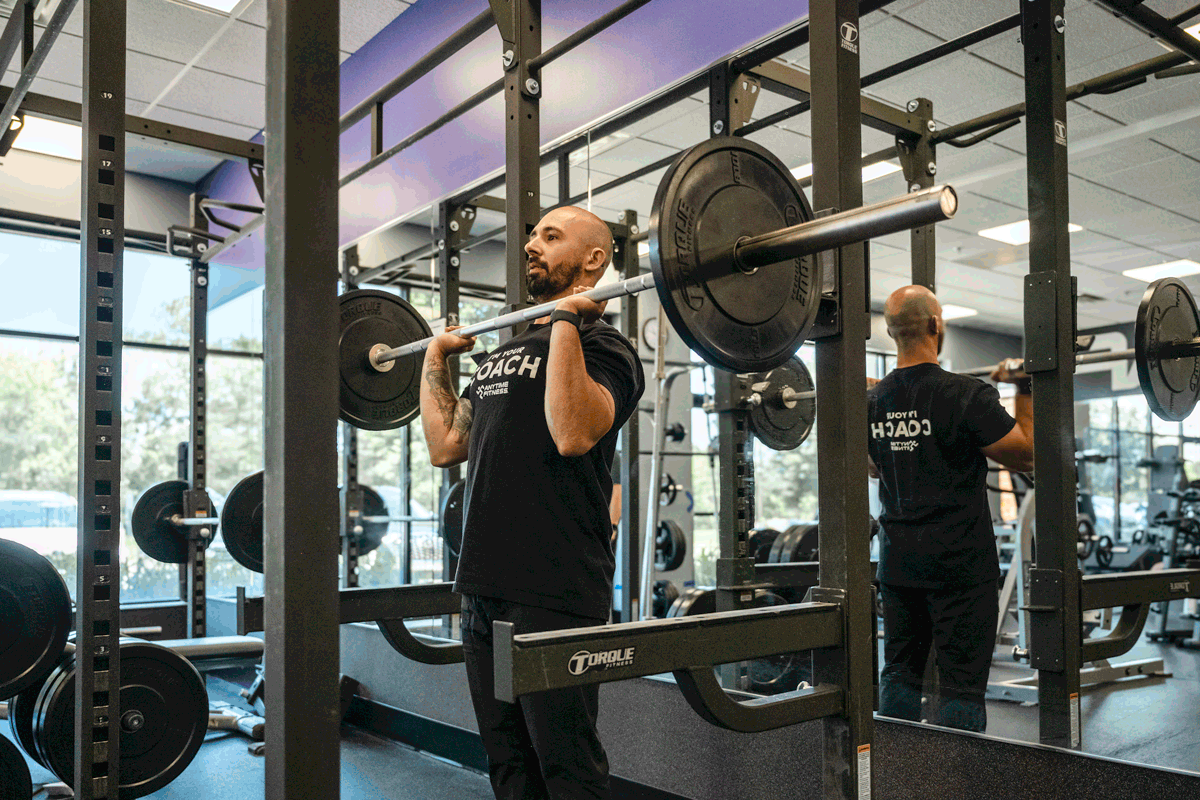 Anytime Fitness coach performing barbell overhead press exercise in a gym environment, emphasizing proper shoulder workout technique.