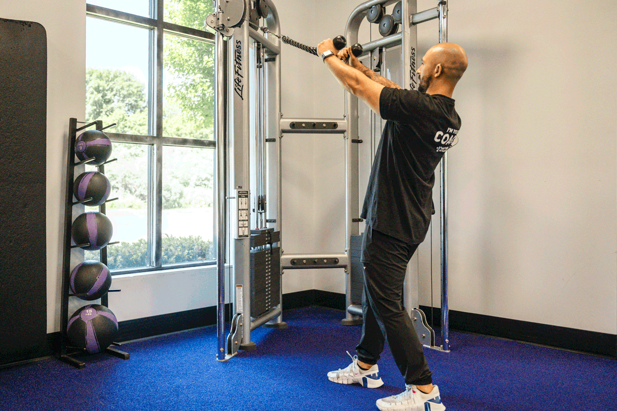 Anytime Fitness coach demonstrating face pull exercise using a cable machine, focusing on shoulder and upper back muscle engagement.