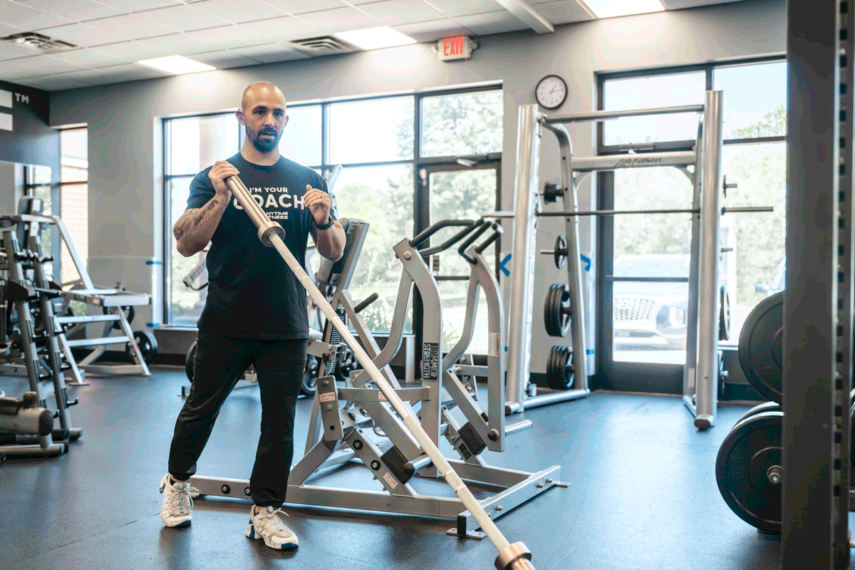Anytime Fitness coach demonstrating landmine press exercise using a barbell, focusing on shoulder and upper body strength.