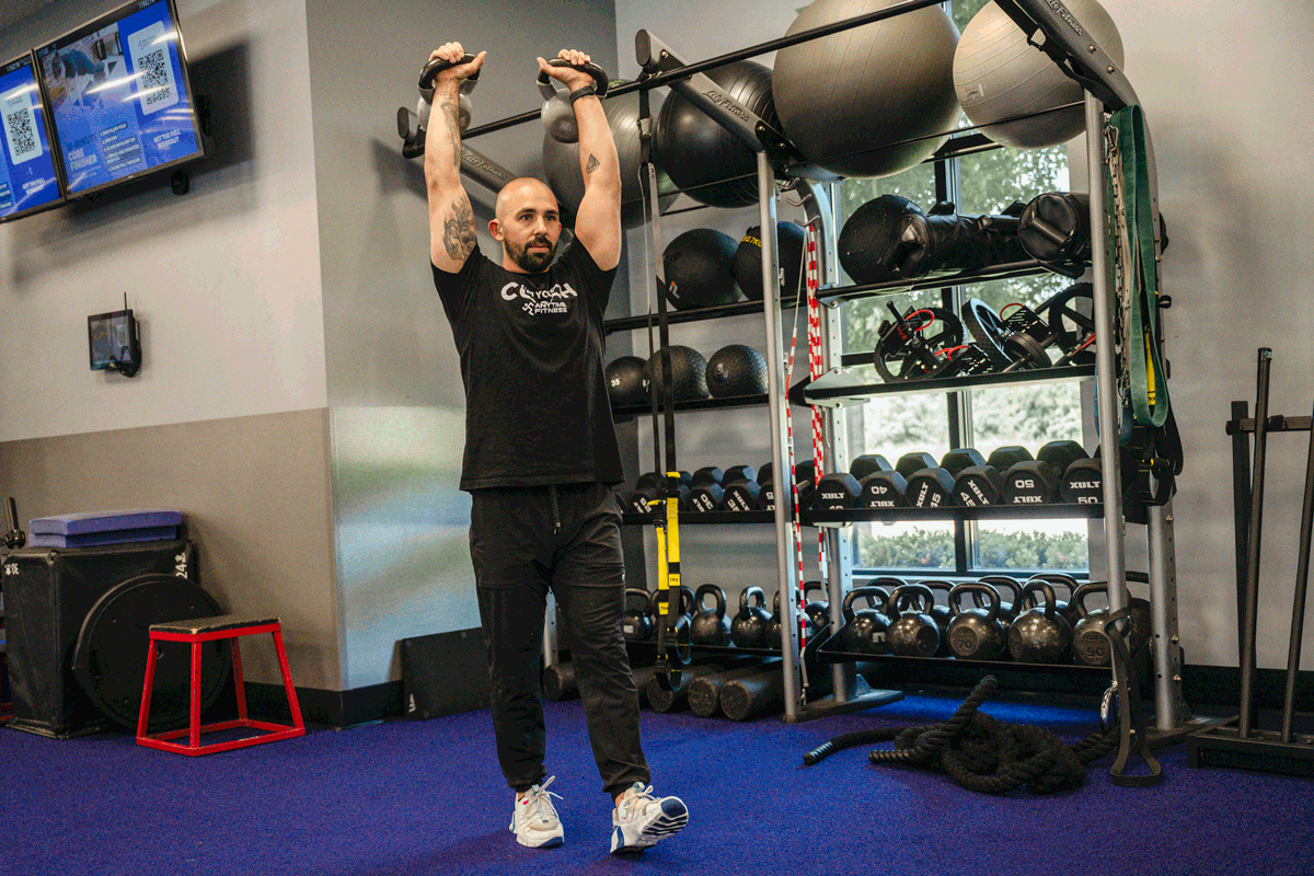 Anytime Fitness coach performing overhead carry exercise with kettlebells, demonstrating full-body strength and stability.