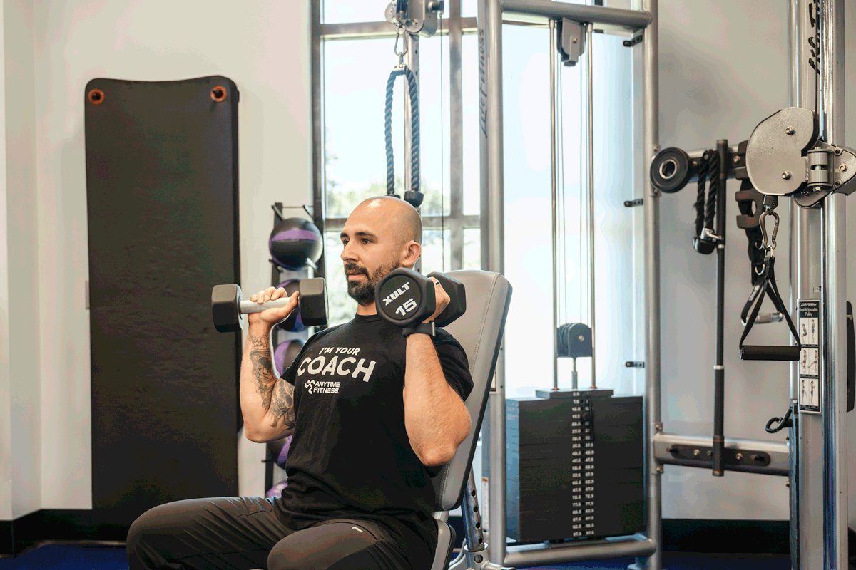 Anytime Fitness coach demonstrating seated overhead press exercise with dumbbells, focusing on shoulder strength and stability in a gym setting.