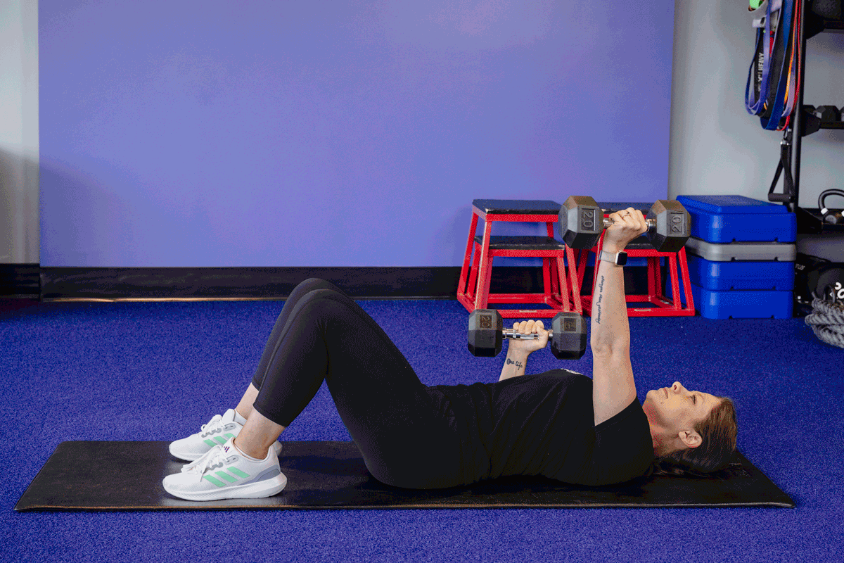 A woman performs an alternating dumbbell floor press exercise, lying on her back with knees bent and feet flat on the mat. She alternates lifting a dumbbell in each hand, engaging her chest and triceps.