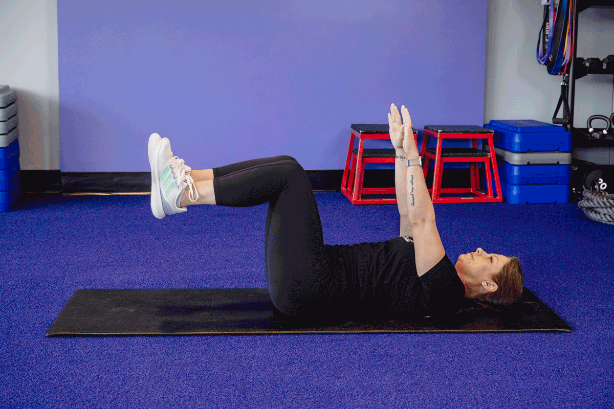 A woman performs a dead bug exercise while lying on her back on a mat. She lifts her arms and legs into the air, moving them in a controlled, alternating manner to engage her core muscles.