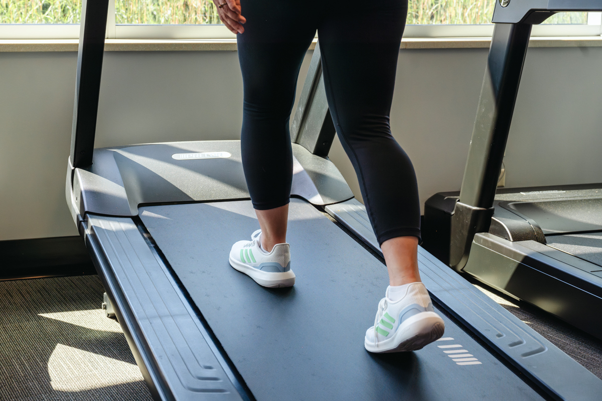 A close-up shot of a woman walking on an incline treadmill, focusing on her lower legs and feet. She wears white and green sneakers, and the gym setting includes large windows that allow natural light to filter in.