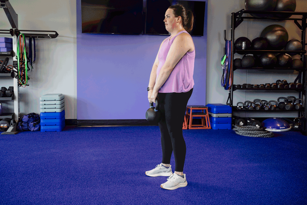 Woman standing in front of a gym equipment wall doing a half deadlift with a kettlebell, focusing on proper form and spine alignment for better posture in a fitness studio.