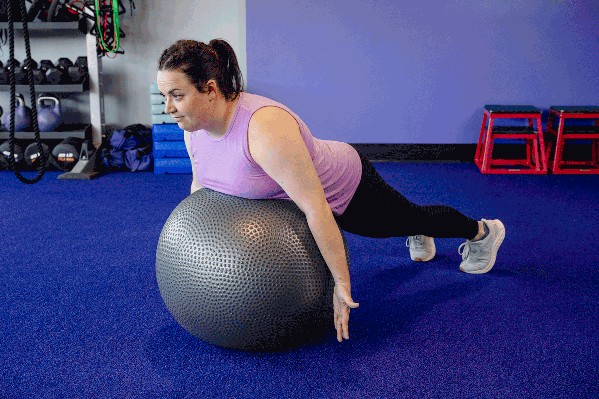 Woman performing a prone middle trapezius exercise with straight legs on a stability ball to improve upper back posture in a fitness studio.