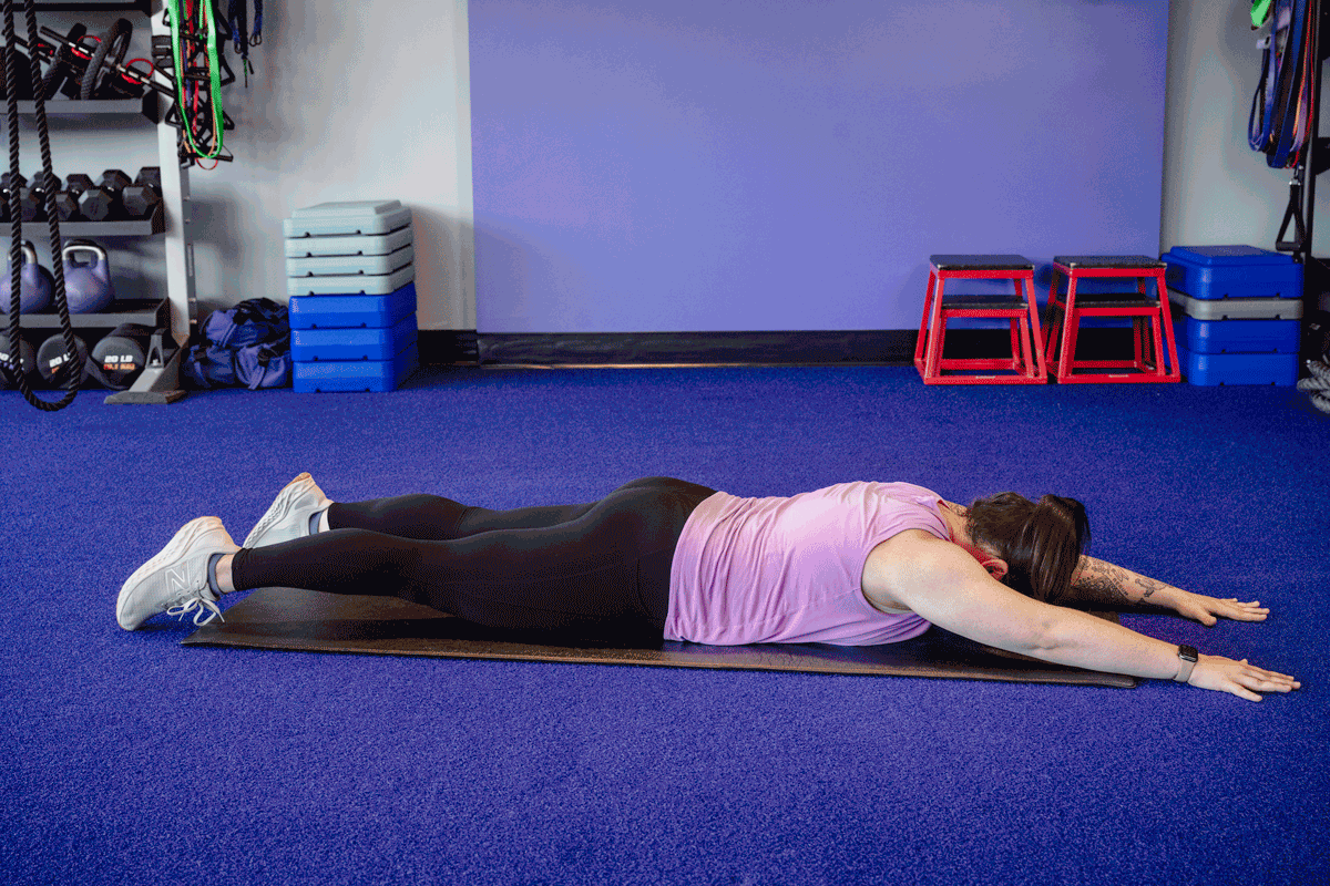 Woman practicing the Superman pose exercise to strengthen lower back and improve posture, lying face down on a gym mat.