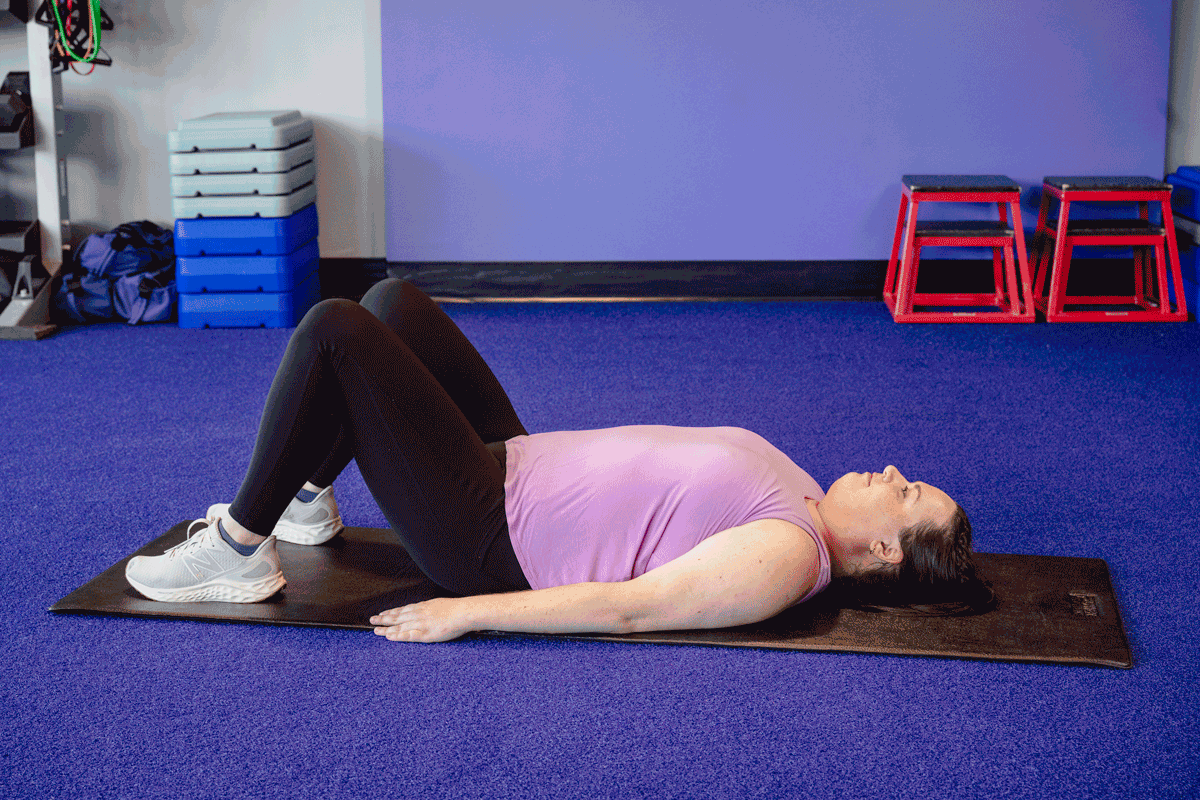 Woman performing a supine bridge exercise to target glutes and core for better posture, lying on a black mat in a fitness studio.