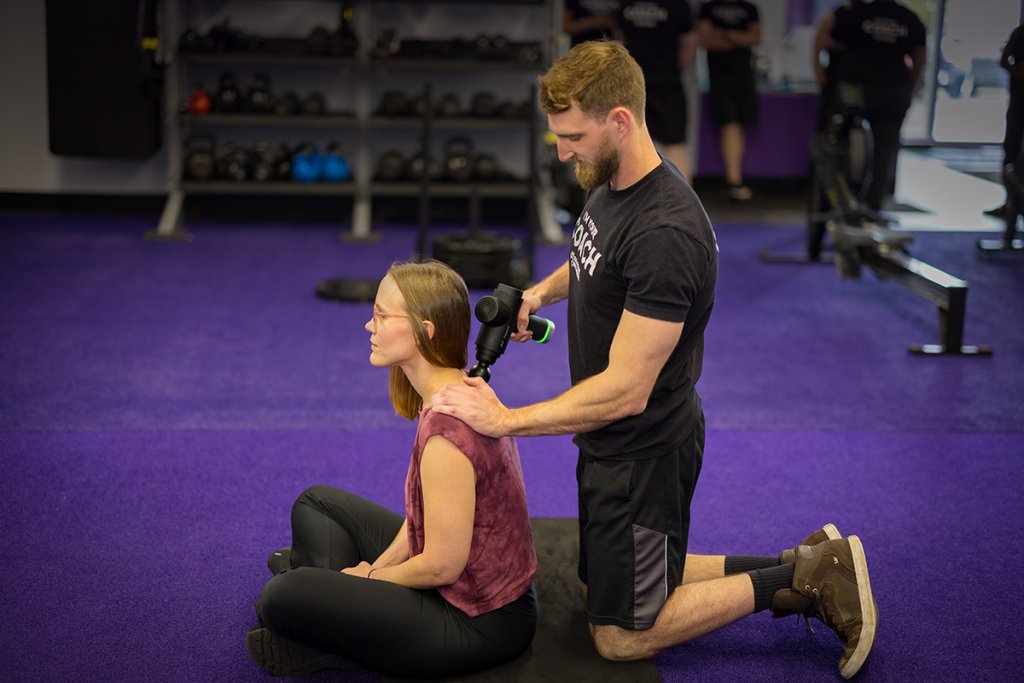 Woman sitting cross-legged on the gym floor while a coach uses a percussion massage tool to relieve tension in her neck and shoulder area.