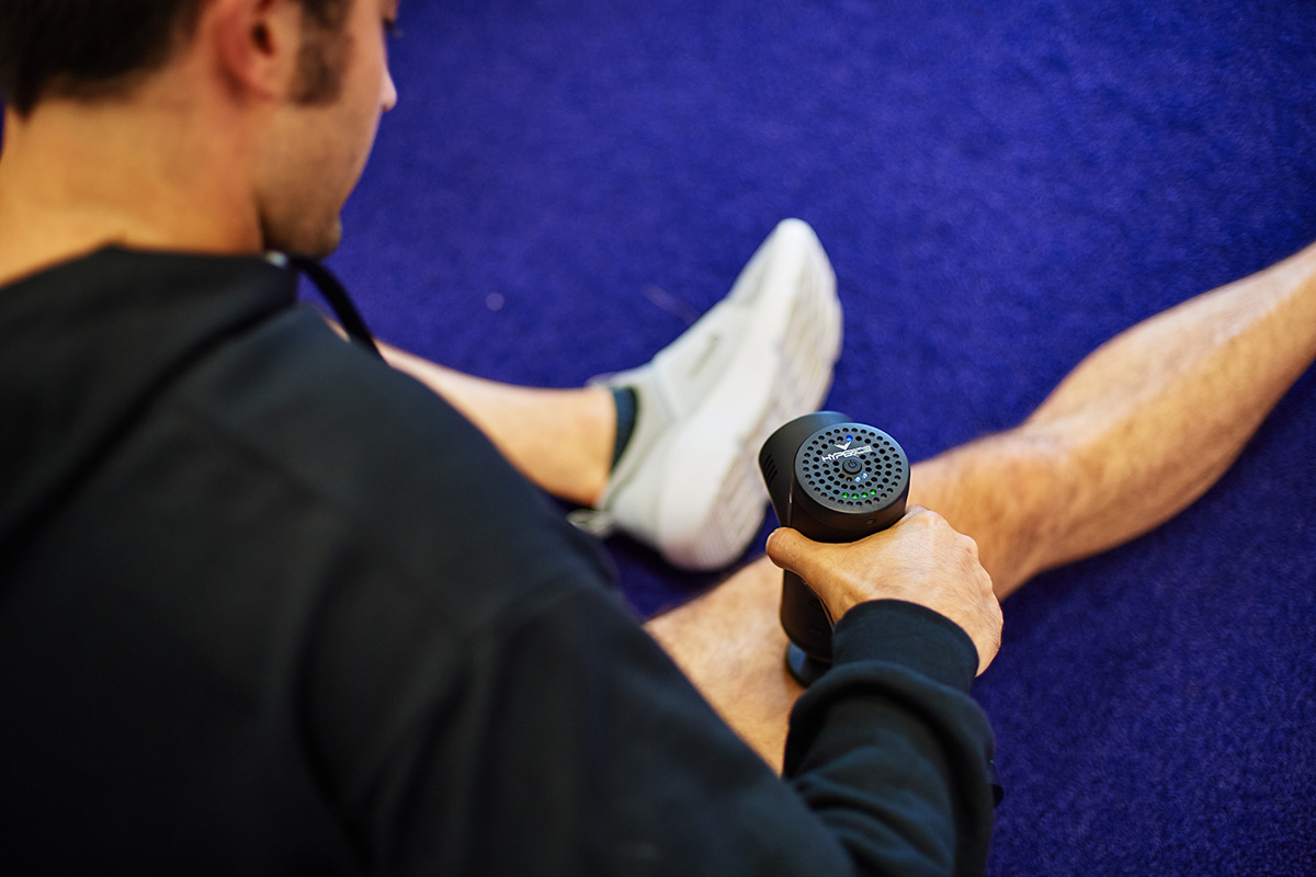 Man sitting on the gym floor holding a percussion massage tool on upper leg for pain relief and relaxation.