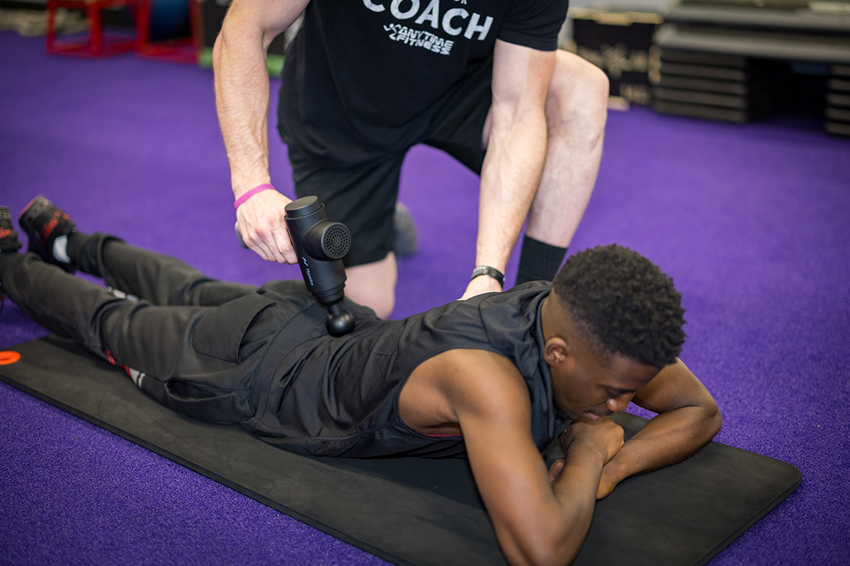 Man lying down on a mat on the gym floor while a coach uses a percussion massage tool on his lower back to reduce muscle tightness.