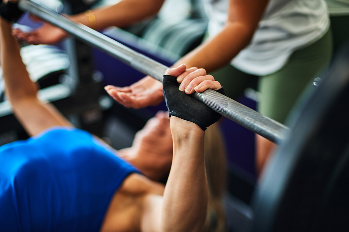 Woman in a blue tank top performing a bench press with a barbell, assisted by a spotter, highlighting proper form and safety in weightlifting.