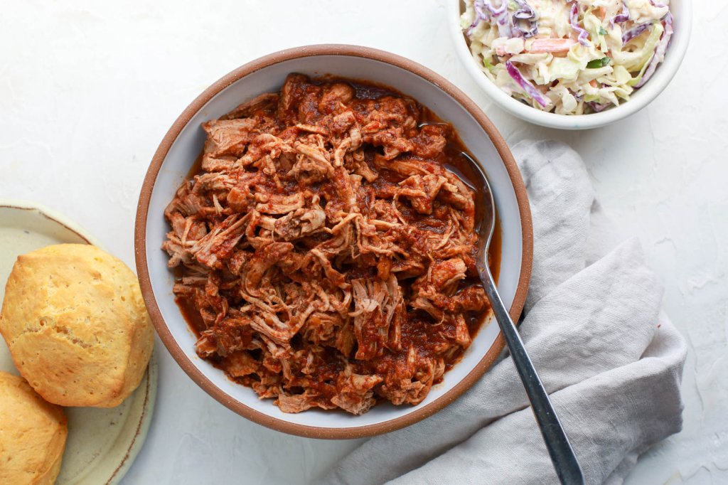 Bowl of shredded raspberry chipotle pork tenderloin, ready to serve with a spoon, next to a bowl of coleslaw and a plate of biscuits.