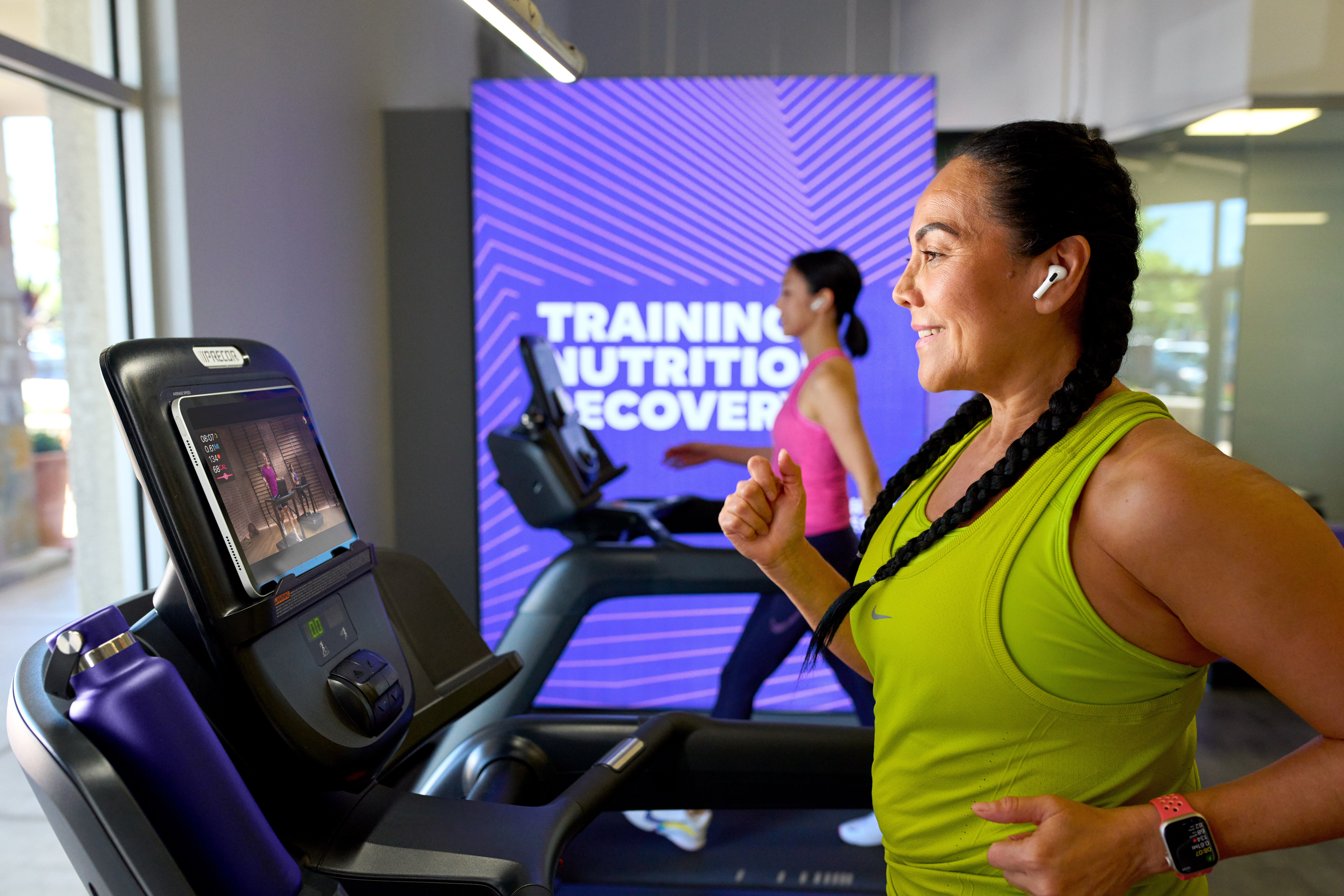 A woman in a bright tank top runs on a treadmill in an Anytime Fitness, following a guided Apple Fitness+ treadmill workout.