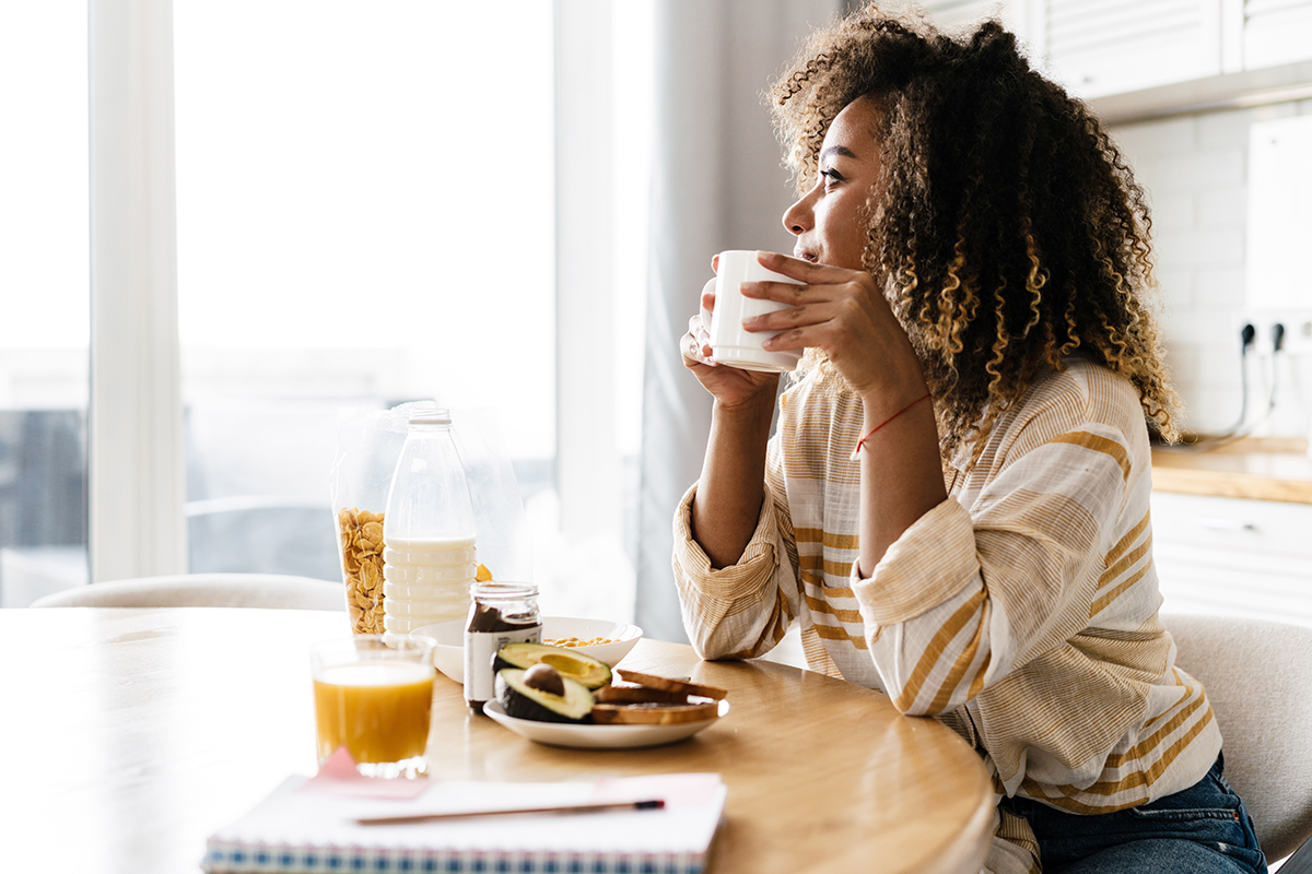 The side view of a smiling woman holding a cup in her hands and looking out the window while sitting in the light kitchen