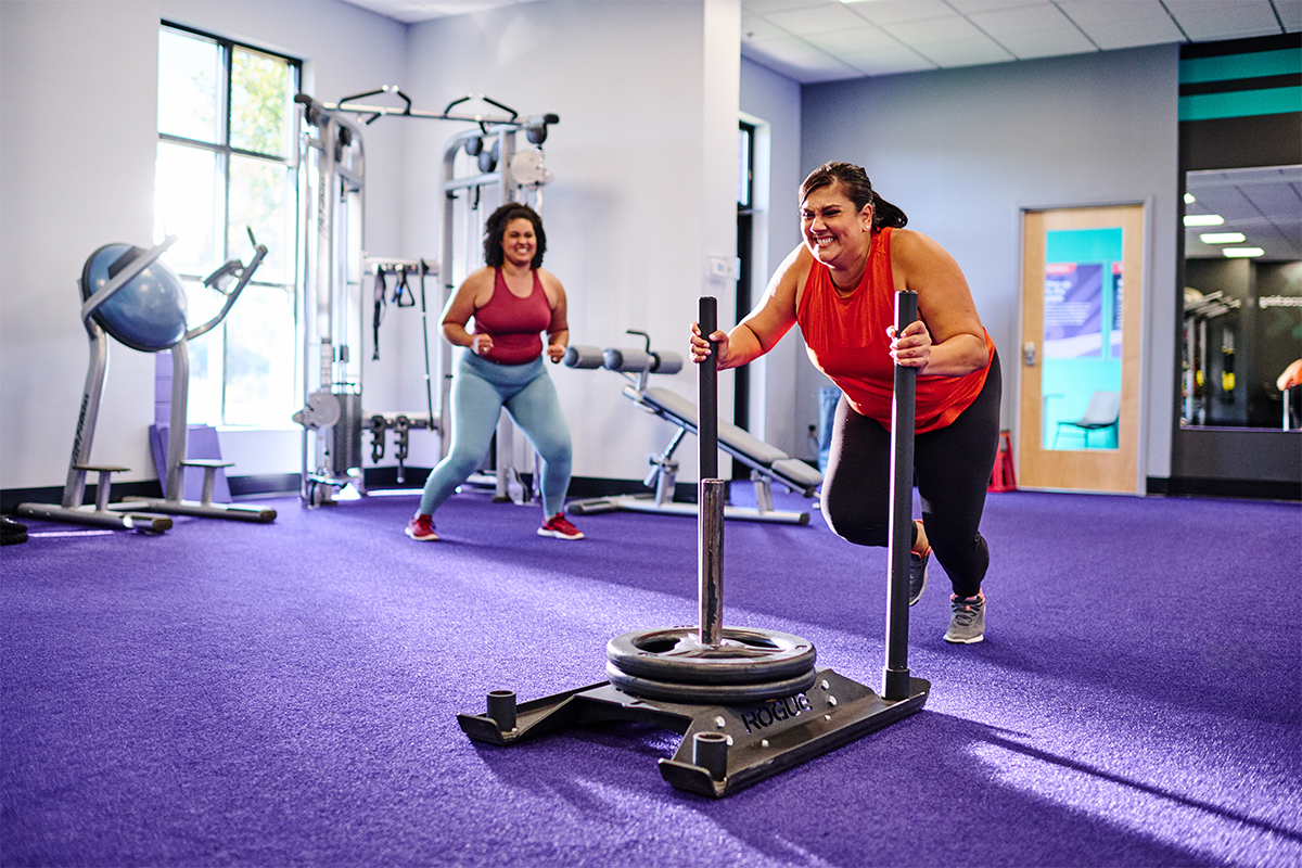 Woman in a gym pushing a sled push while a friend encourages her from the side.