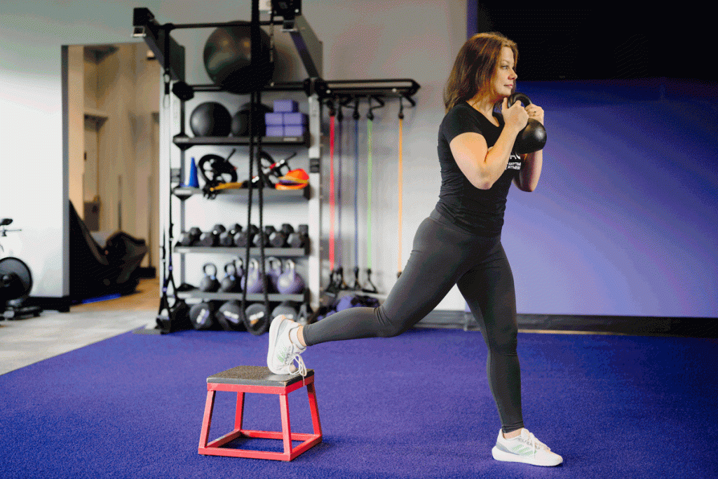 Woman performing a kettlebell goblet Bulgarian split squat using a box in a gym setting.