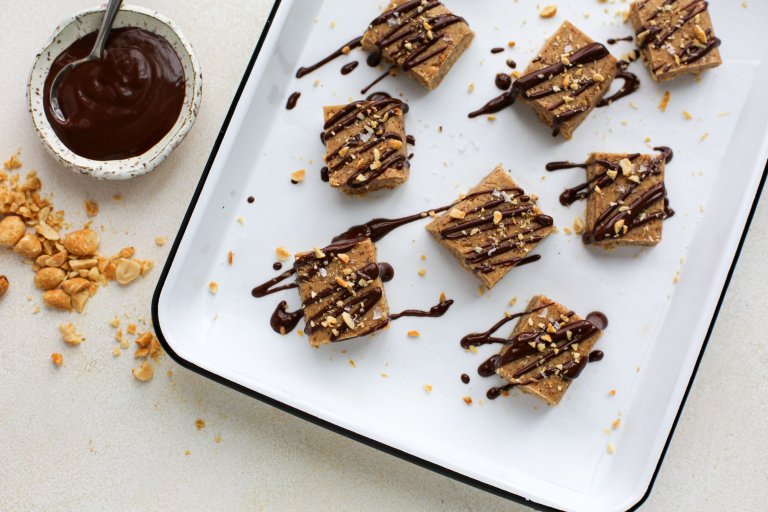 Tray of protein bites prepared with nuts and drizzled chocolate on top, cut into squares with additional toppings on the side.