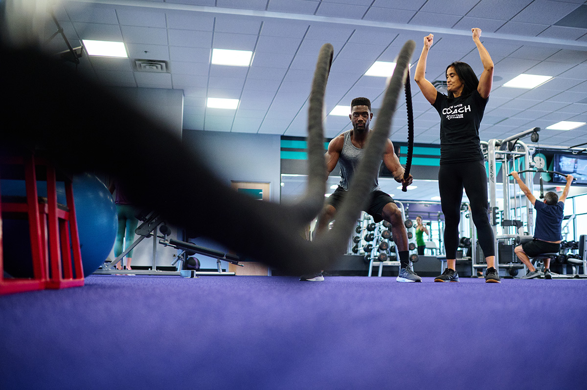 A man performing an intense battle rope exercise with a Coach cheering him on in a gym, promoting strength, endurance, and teamwork.