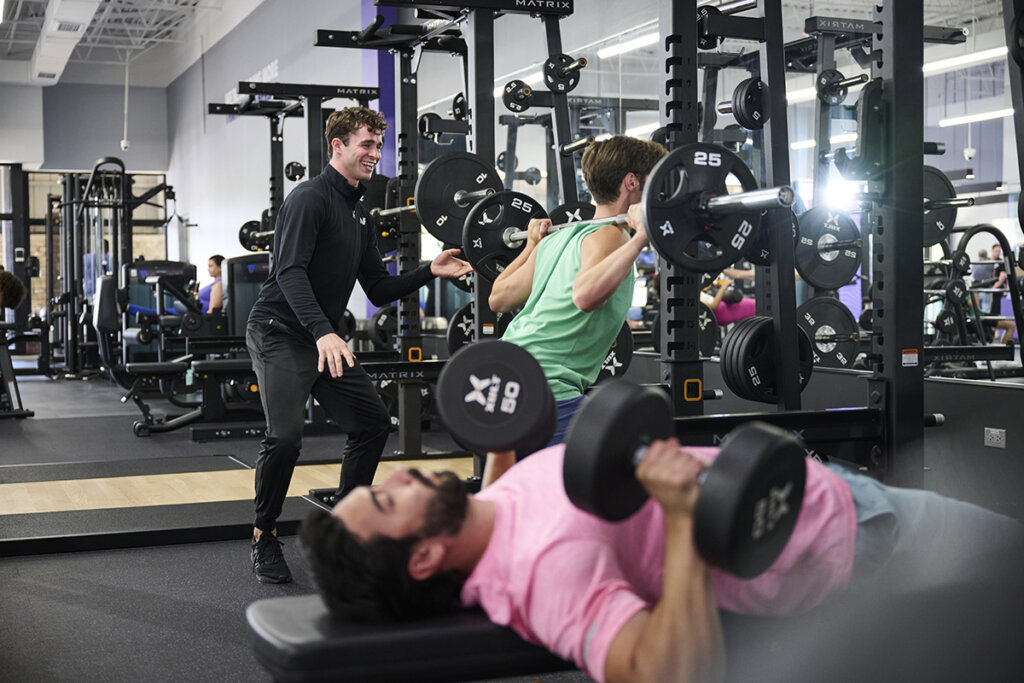 A gym session featuring a trainer coaching a client during a squat with a barbell, while another individual performs dumbbell presses, highlighting teamwork and individualized training.