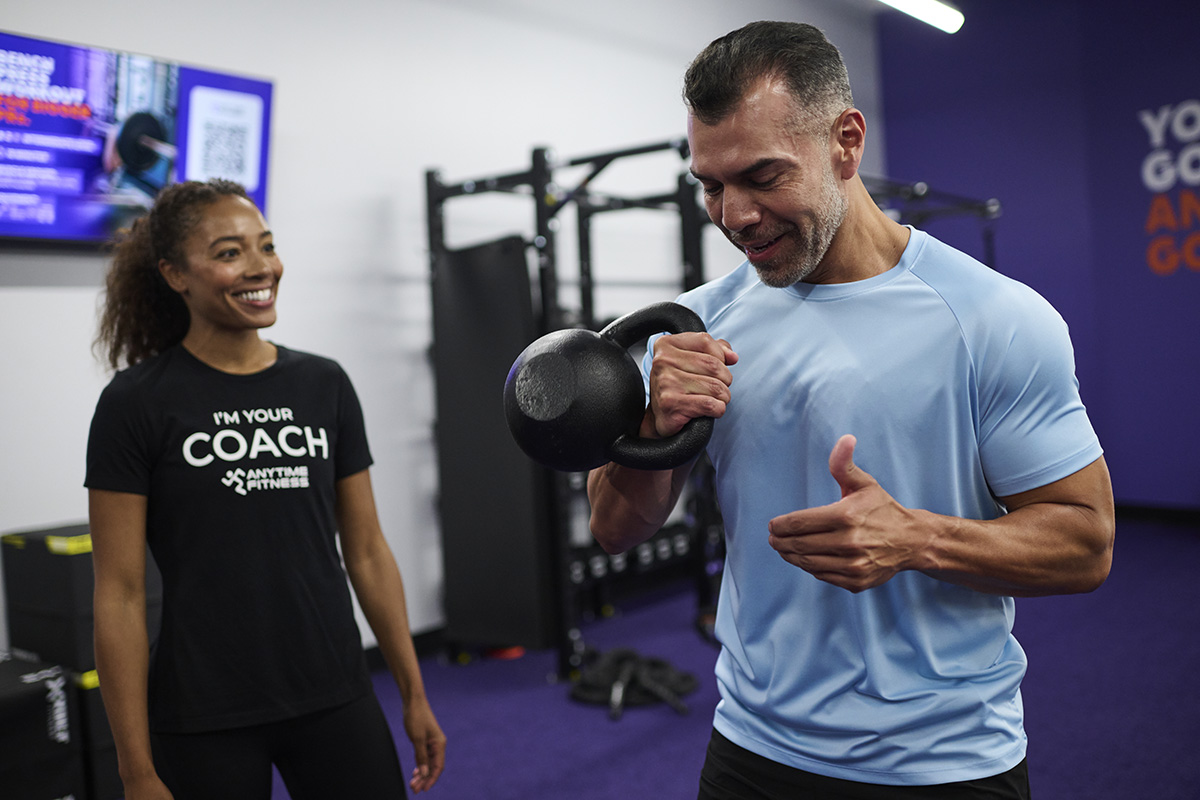 A man in a blue shirt doing a kettlebell curl while a fitness coach in a black "I'm Your Coach" shirt provides guidance and encouragement.