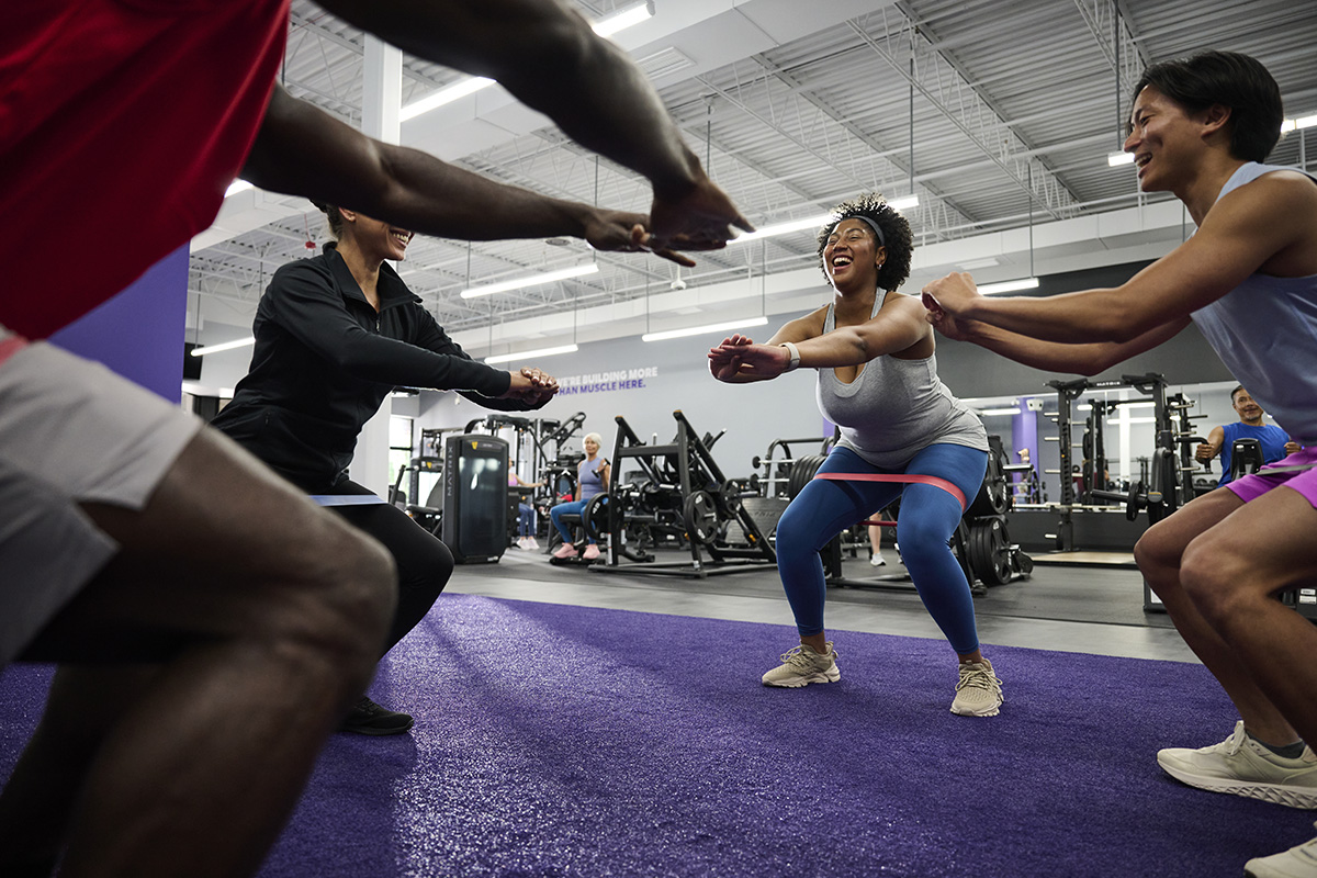 A group of individuals performing squats with resistance bands in a gym, led by an instructor, promoting teamwork and functional strength training.