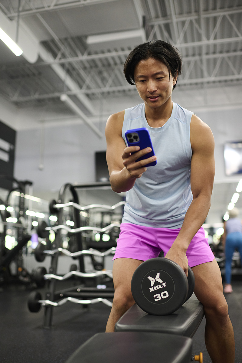 A man in a light gray tank top and pink shorts checking his smartphone while holding a 30-pound dumbbell on a bench, emphasizing fitness tracking and multitasking.