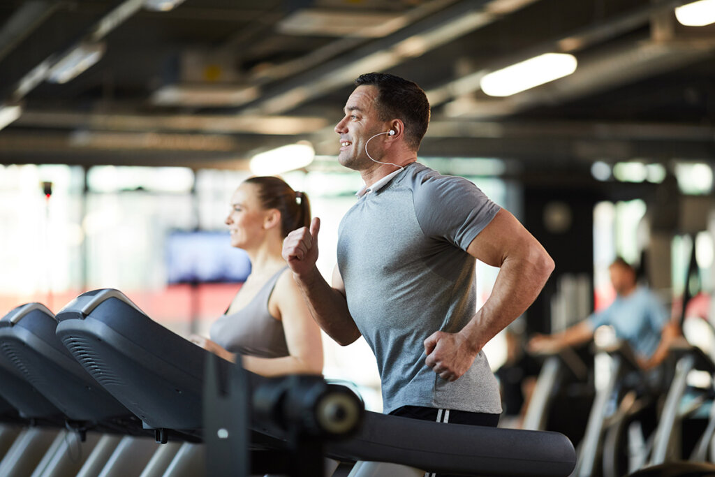 A man focused on his treadmill run, wearing earbuds, and training in a bustling fitness center.