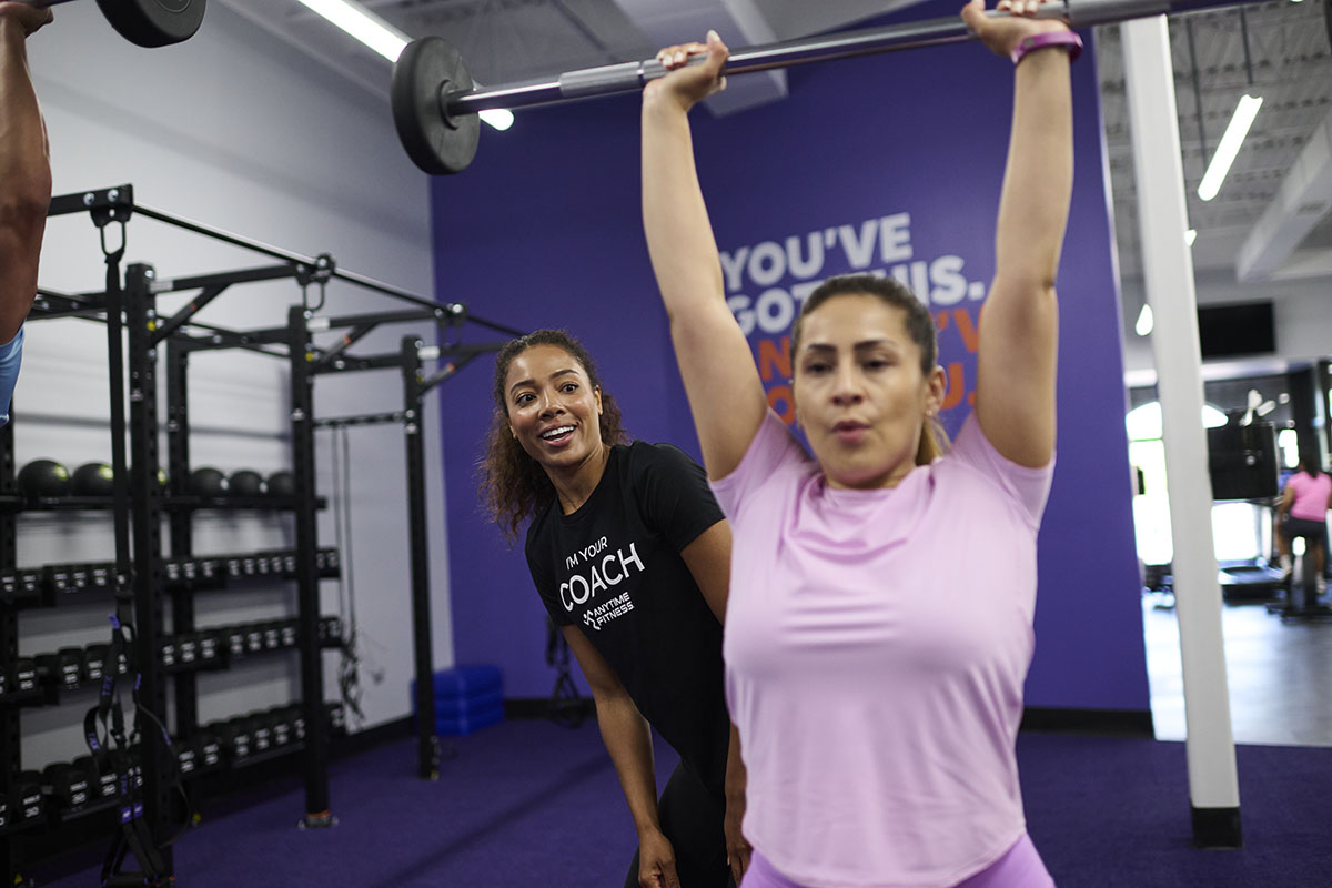 A Coach cheering on a woman as she lifts a barbell overhead, showcasing the value of supportive Coaching during strength training.