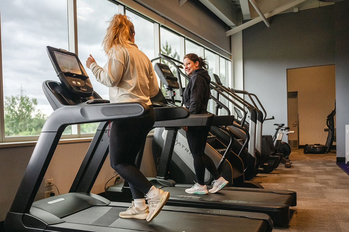 Two women enjoying a treadmill workout in a bright gym, illustrating the benefits of exercising with a friend.