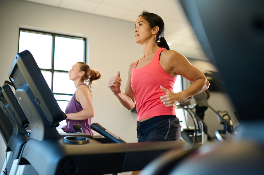 Two women in colorful tank tops do a treadmill workout in an Anytime Fitness gym.