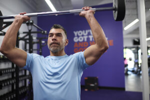 Man in an Anytime Fitness gym engaging in a muscle-building workout, raising a weighted barbell overhead.