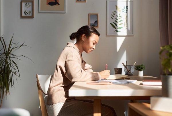 Young woman writing in a notebook at home with sun rays shining.